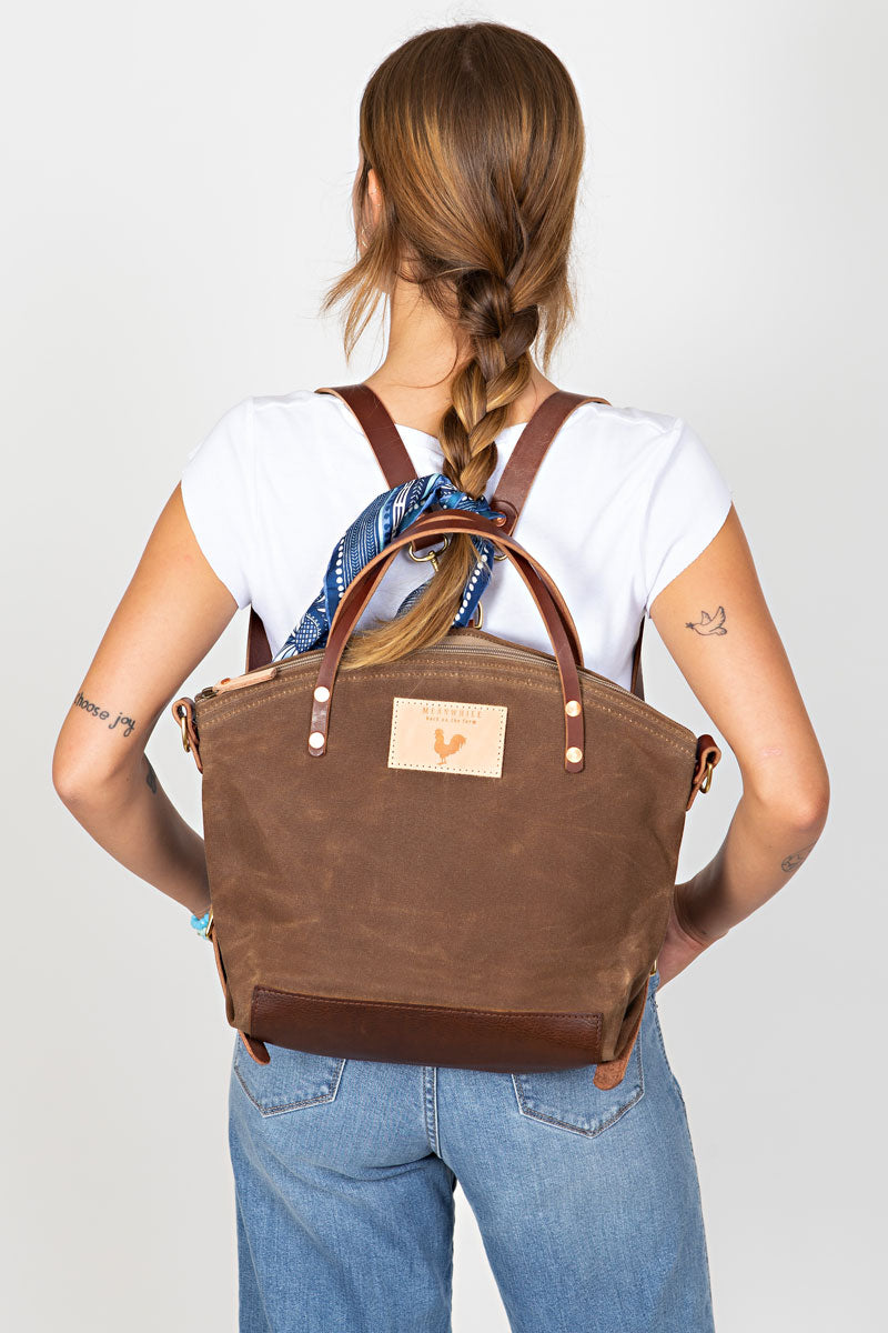 A woman wearing the field tan backpack with brown straps and the meanwhile logo.