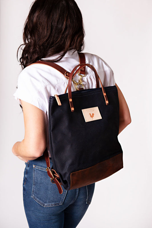 A woman wearing the navy backpack with dark brown straps and the meanwhile logo.