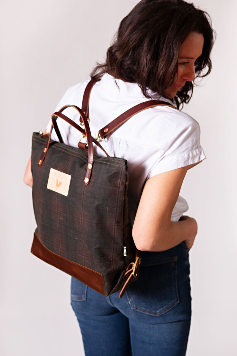 Woman wearing a green plaid backpack with meanwhile logo and dark brown straps.