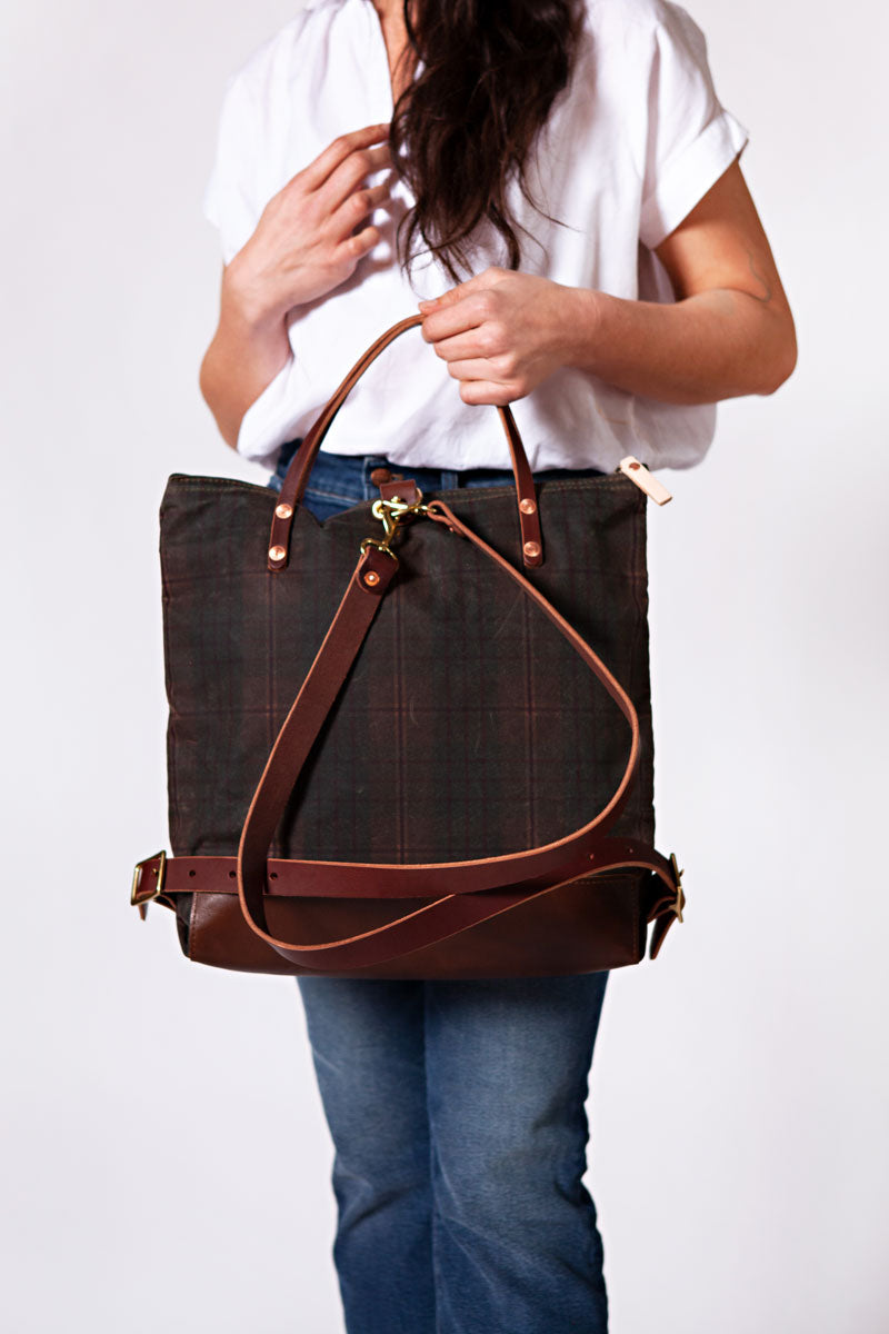 Woman holding a green plaid backpack with meanwhile logo and dark brown straps.