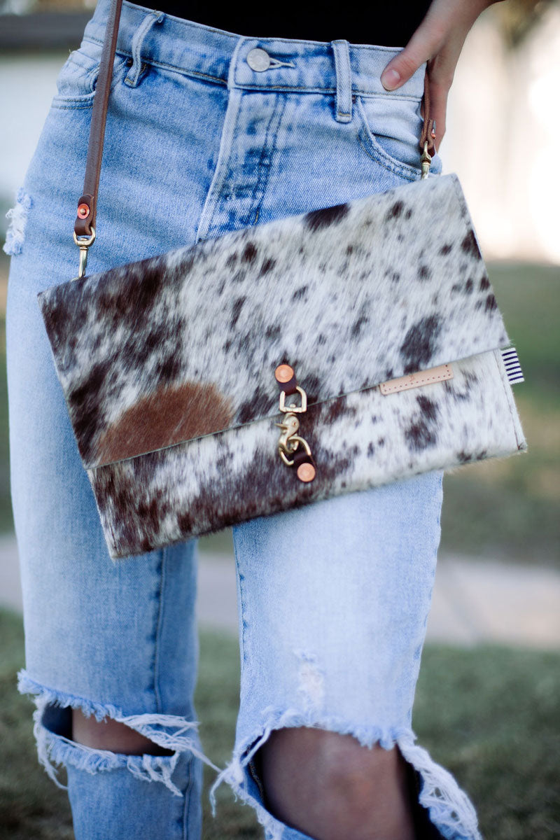 Picture of model in ripped jeans wearing a large fold over purse in a brushed cowhide leather. Bag has brown adjustable leather strap.