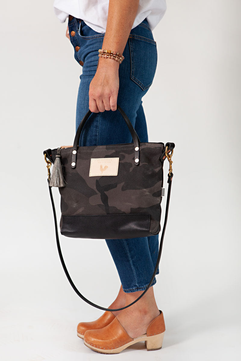 A woman holding the graphite camouflage small tote bag with a grey tassel and the meanwhile logo.