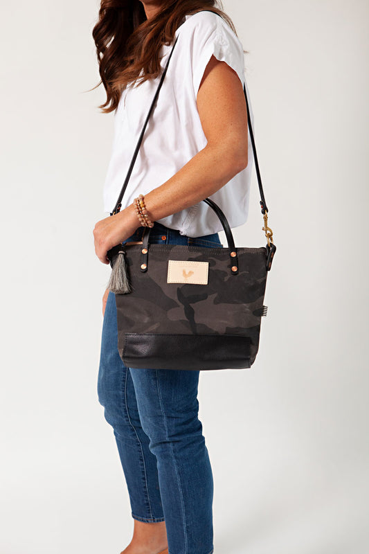 A woman holding the graphite camouflage small tote bag with a grey tassel and the meanwhile logo.