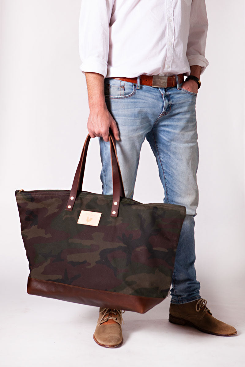 A man holding the camouflage weekend bag with the meanwhile logo and dark brown shoulder straps.
