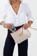 Woman placing her phone in the white and tan clutch with a light brown strap, tan tassel, and a bright pink zipper. 